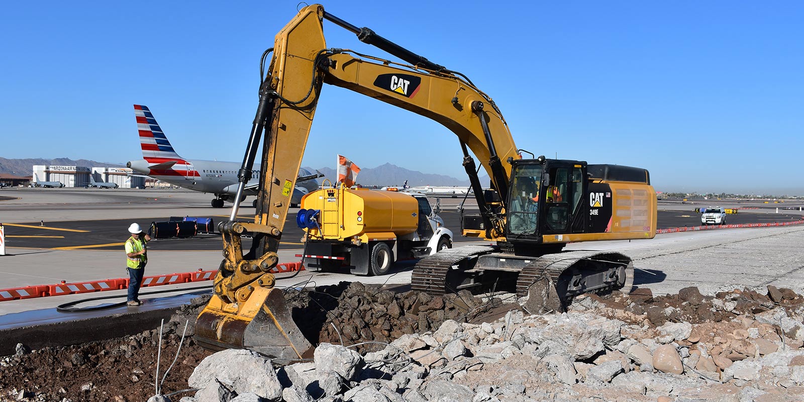 Excavator breaking out concrete pavement on an airport apron, with an airliner at the terminal behind
