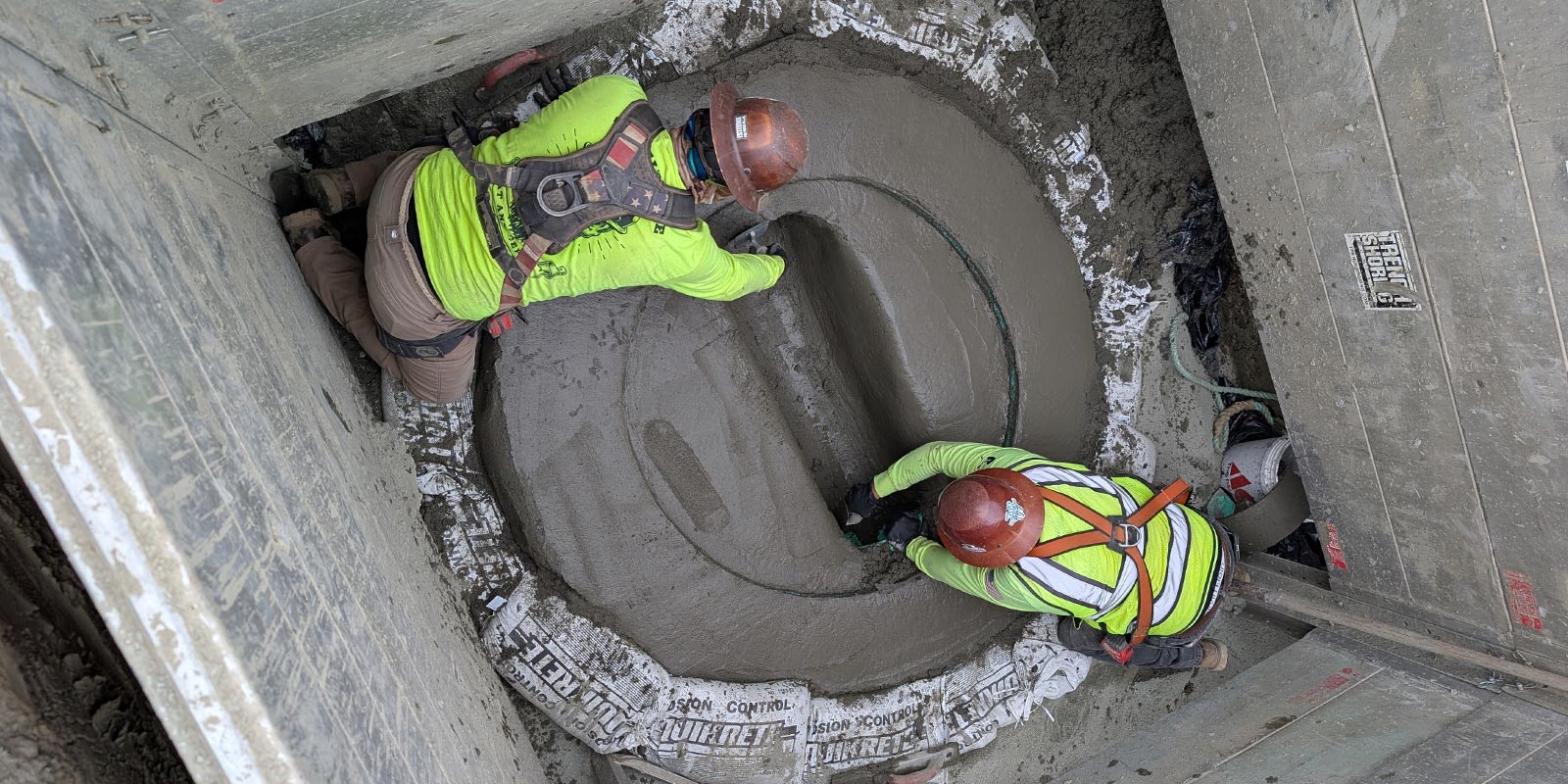 Two workers in high-visibility gear finishing concrete inside a shored trench
