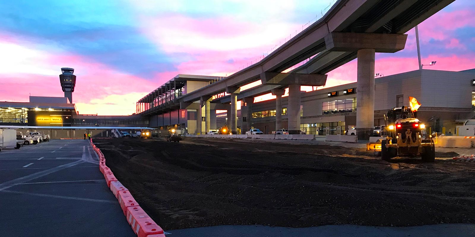 Grader working on airport terminal roadway at dusk beneath an elevated sky train guideway
